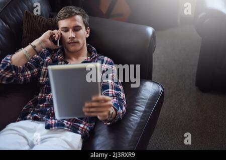 Klar. Ich melde mich gerade an. High-Angle-Aufnahme eines hübschen jungen Mannes, der sein Tablet und Handy benutzt, während er zu Hause auf dem Sofa sitzt. Stockfoto