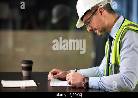 Füllen Sie einige Papiere aus. Eine kurze Aufnahme eines hübschen, reifen männlichen Bauarbeiters, der die Papiere an seinem Schreibtisch im Büro ausfüllte. Stockfoto