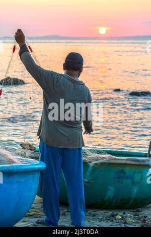 Blick auf den Strand am Korbbootdock bei Sonnenuntergang, während sich die Fischer an ihren Netzen darauf vorbereiten, am nächsten Morgen in Mui Ne, Vietnam, die Segel zu setzen Stockfoto