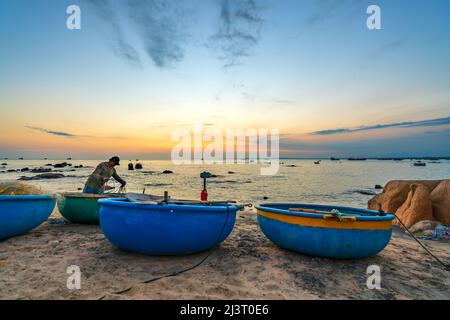 Blick auf den Strand am Korbbootdock bei Sonnenuntergang, während sich die Fischer an ihren Netzen darauf vorbereiten, am nächsten Morgen in Mui Ne, Vietnam, die Segel zu setzen Stockfoto