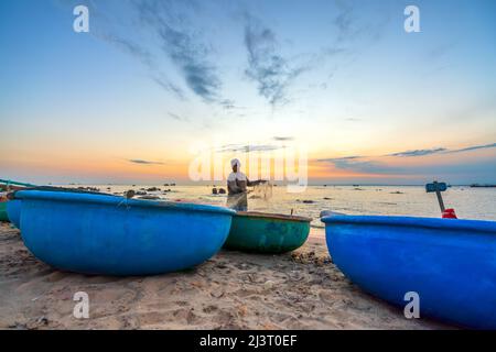 Blick auf den Strand am Korbbootdock bei Sonnenuntergang, während sich die Fischer an ihren Netzen darauf vorbereiten, am nächsten Morgen in Mui Ne, Vietnam, die Segel zu setzen Stockfoto