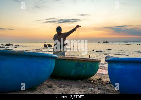 Blick auf den Strand am Korbbootdock bei Sonnenuntergang, während sich die Fischer an ihren Netzen darauf vorbereiten, am nächsten Morgen in Mui Ne, Vietnam, die Segel zu setzen Stockfoto