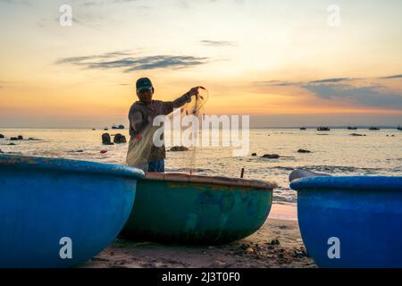 Blick auf den Strand am Korbbootdock bei Sonnenuntergang, während sich die Fischer an ihren Netzen darauf vorbereiten, am nächsten Morgen in Mui Ne, Vietnam, die Segel zu setzen Stockfoto