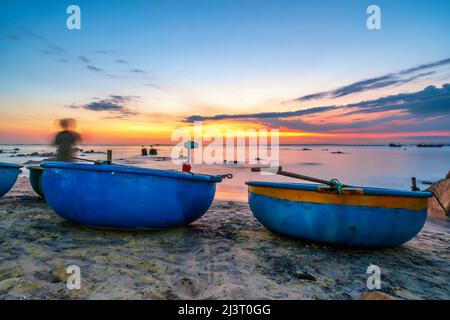 Blick auf den Strand am Korbbootdock bei Sonnenuntergang, während sich die Fischer an ihren Netzen darauf vorbereiten, am nächsten Morgen in Mui Ne, Vietnam, die Segel zu setzen Stockfoto