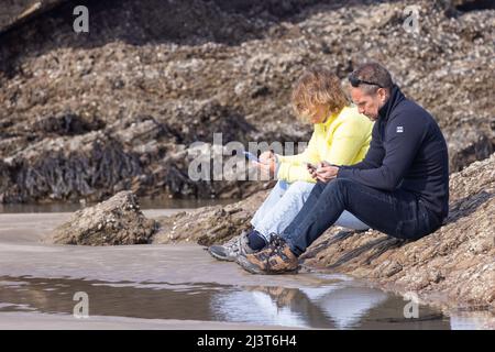 Zwei Menschen, die bei Ebbe auf den Felsen sitzen, konzentrieren sich beide auf ihre Mobiltelefone Stockfoto