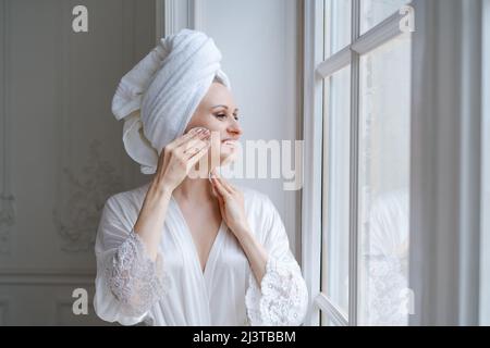 Glückliche Frau nach dem Duschen kümmert sich um saubere Haut und Poren, hält Wattepad Phase Cleanser entfernt Make-up und Schmutz genießt eine effektive Beauty-Behandlung, steht am Fenster mit Bademantel und Handtuch auf dem Kopf Stockfoto