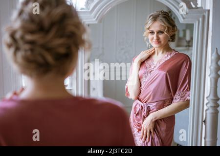 Eine glückliche Frau sieht ihre Spiegelung in einem Spiegel in rosa Seidengewand am Fenster am Morgen an. Schönes Make-up und Haare. Haut- und Körperpflege zu Hause. Schönheit und Gesundheit von Frauen Stockfoto