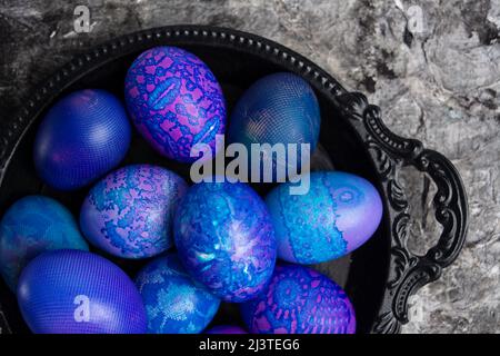 Ostereier. Blau-lila Ostereier mit Spitze bemalt, in einer schwarzen Schale auf einem schwarzen rustikalen Hintergrund, Draufsicht. Ei Hintergrund Muster. Stockfoto