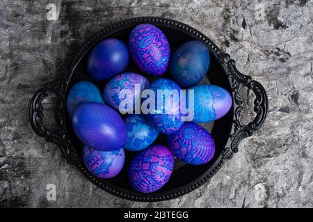 Ostereier. Blau-lila Ostereier mit Spitze bemalt, in einer schwarzen Schale auf einem schwarzen rustikalen Hintergrund, Draufsicht. Ei Hintergrund Muster. Stockfoto