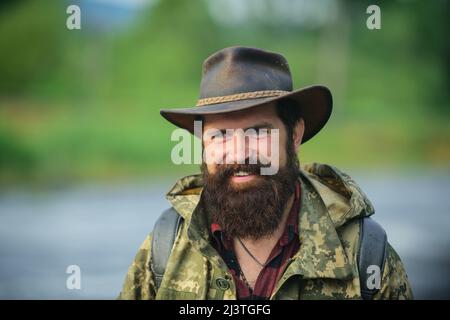 Porträt eines Reisenden bärtigen Mannes in Cowboyhut. Nahaufnahme eines glücklichen Mannes mittleren Alters, der auf dem Land lächelt. Stockfoto