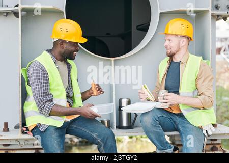 Zwei Ingenieure, die Harthüte und Sicherheitswesten tragen, sitzen auf einem Zementbalken und ruhen sich während der Pause auf der Baustelle aus Stockfoto