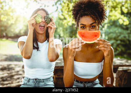 Fröhlichen multikulturellen jungen erwachsenen Freundinnen sitzen auf dem Land unter den Bäumen essen frische Wassermelone und verstecken die Gesichter mit Scheiben und Schalen - g Stockfoto