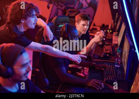 Junger Mann, der auf den Computerbildschirm zeigt, während er dem afroamerikanischen Spieler Ratschläge gibt, der sich auf das Spiel im eSports Club konzentriert Stockfoto