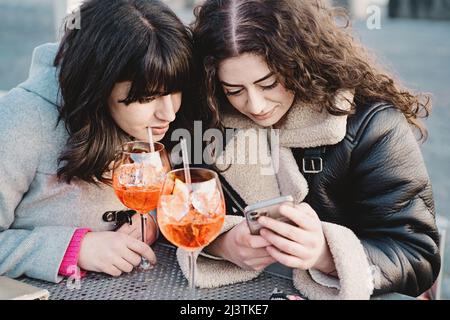 Zwei junge Frauen checken auf ihren Smartphones soziale Netzwerke, während sie zwei Spritz-Cocktails am Kaffeetisch trinken Stockfoto