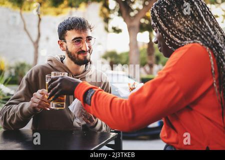 Zwei junge Leute, die im Café sitzen und eine Pause machen - ein multikulturelles junges Paar, das mit Bier anstoßen und Sandwiches essen kann Stockfoto