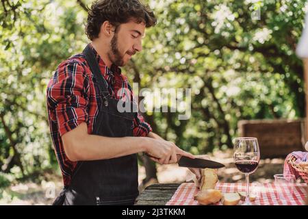 Bärtiger Hipster-Mann, der auf dem Land das Brot über einem Tisch schneidet - junger Mann, der in der Natur Essen für das Picknick zubereitet Stockfoto