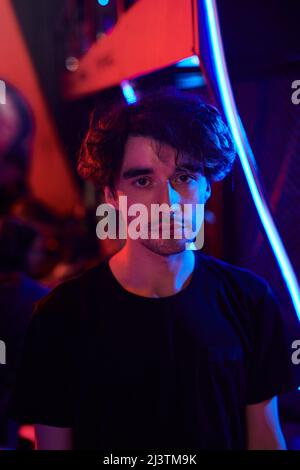 Porträt eines traurigen jungen kaukasischen Mannes in schwarzem T-Shirt, der in der eSports Bar, blauen und roten Lichtern steht Stockfoto