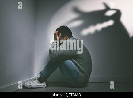 Wenn Träume wahr werden, können auch Alpträume wahr werden. Aufnahme eines jungen Mannes, der in der Ecke eines dunklen Raumes mit einer unheimlichen Figur an der Wand sitzt. Stockfoto