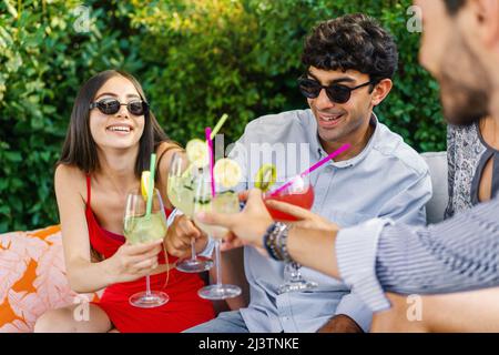 Fröhliche Freunde, die im Freien auf einer Couch tropische Fruchtcocktails toasten - Junge Leute, die in der Happy Hour gerne mit Gläsern klirren. Fokus Stockfoto