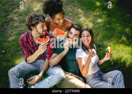 Multikulturelle Gruppe junger Menschen, die Wassermelone essen und auf dem Rasen auf dem Land sitzen Stockfoto