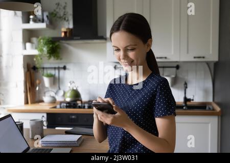 Lächelnde junge attraktive Frau, die in der Küche mit Handy steht Stockfoto