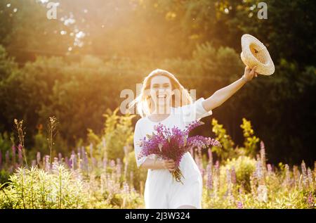 Junge Blondine mit kurzen Haaren in einem sommerlichen weißen Kleid. Frau posiert in einem Lupinenfeld Stockfoto