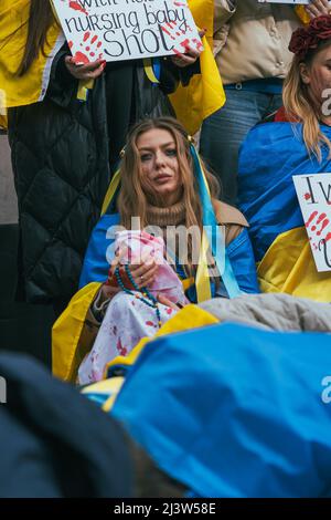 Eine pro-ukrainische Frau sitzt auf den Straßen einer Demonstration mit einer ukrainischen Flagge um ihren Körper und hält ein Baby, das mit einem Blatt Kriegsblut bedeckt ist Stockfoto