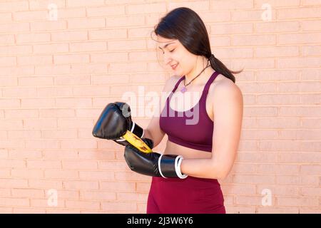 Eine junge kaukasische Frau trägt schwarze Boxhandschuhe und versucht, eine Banane zu schälen. Im Hintergrund ist eine Ziegelmauer. Stockfoto