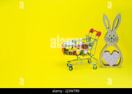 Osterhase aus Holz und Warenkorb mit Süßigkeiten auf gelbem Hintergrund Stockfoto