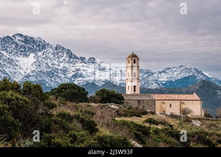 Kirche im Dorf Sant'Antonino in der Balagne auf Korsika mit schneebedeckten Bergen in der Ferne Stockfoto