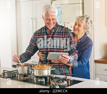 Was kocht, gut aussehend. Eine kurze Aufnahme eines älteren Ehepaares, das ein digitales Tablet verwendet, während es zu Hause in der Küche zusammen kocht. Stockfoto