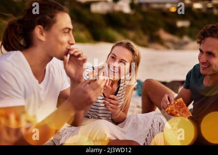 Pizza ist gutes Essen. Aufnahme einer Gruppe von Freunden, die zusammen am Strand sitzen. Stockfoto