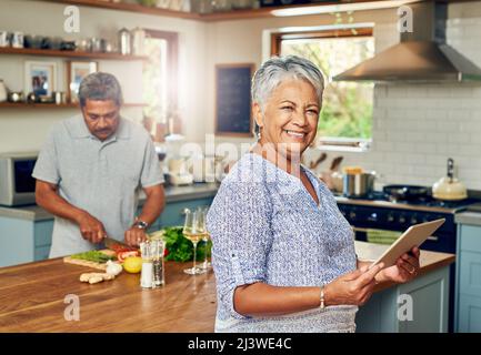 Der intelligenteste kleine Soux-Koch, den es gibt. Porträt einer reifen Frau, die ein digitales Tablet benutzt, während sie zu Hause mit ihrem Mann eine Mahlzeit zubereitet. Stockfoto