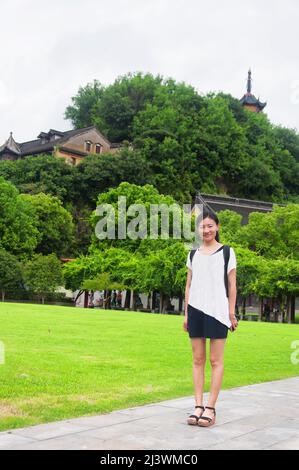 Eine chinesische Frau, die am Rande eines Rasens steht und die Cishou-Pagode über den Bäumen im malerischen Gebiet des Jinshan-buddhistischen Tempels in Zhenjiang China aufsteigt Stockfoto