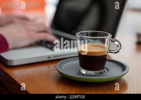 Frischer Espresso-Kaffee auf einem Holzschreibtisch aus der Nähe. Verwischen Sie am Morgen im Büro die Hände von Frauen auf einer Laptop-Tastatur Stockfoto