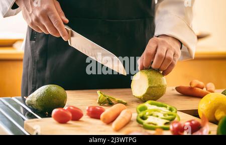 Koch mit schwarzer Schürze, die Zucchini auf einem Schneidebrett schneidet, Hand kochen und ein Messer aus nächster Nähe halten. Frisches Gemüse auf Küchentisch, Restaurant vegetaria Stockfoto