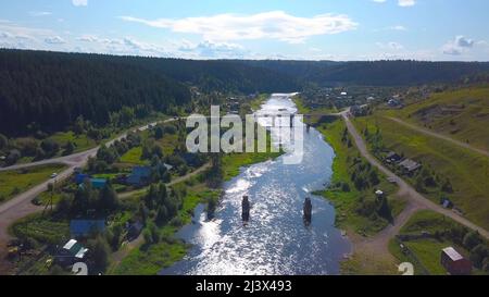 Eine Luftdrohnenlandschaft eines kleinen Dorfes mit Fluss und grünen bewaldeten Bergen. Clip. Eine Brücke über zwei Flussufer auf bewölktem Himmel Hintergrund Stockfoto