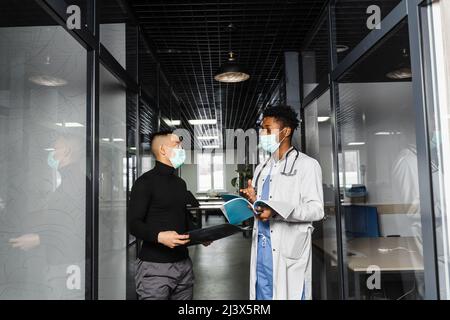 Diskussion und Beratung zwischen schwarzem Arzt und Patient in der medizinischen Klinik. Afrikanischer Chirurg behandelt asiatischen gutaussehenden Mann Stockfoto