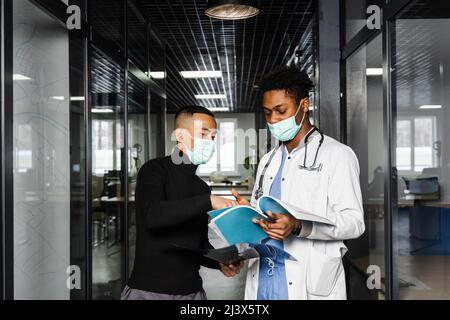 Diskussion und Beratung zwischen schwarzem Arzt und Patient in der medizinischen Klinik. Afrikanischer Chirurg behandelt asiatischen gutaussehenden Mann Stockfoto