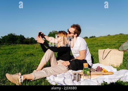 Soziales junges Paar, das während eines Picknicks auf einem frühlingsgrassfield auf dem Land ein Selfie gemacht hat. An einem sonnigen Tag auf einer Decke sitzen. Stockfoto