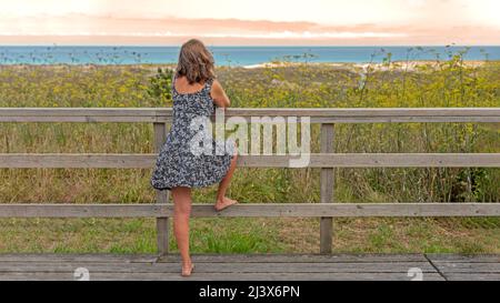 Blonde Frau auf Holzsteg oder Brücke mit fließenden Kleid vom Wind bewegt und modisch im Meer Stockfoto