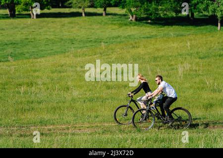 Lange Aufnahme eines Paares, das mit dem Fahrrad auf dem Land unterwegs ist. Seitenansicht. Blick von hinten. Reiten auf der Strecke entlang von lebhaften Grasfeldern. Stockfoto