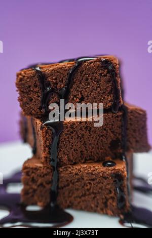 Sehr nahe Aufnahme eines Stapels von Schokoladenbrownies mit Schokoladensträngen, die auf einen weißen Teller mit violettem Hintergrund fallen. Stockfoto