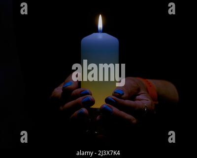 Eine brennende Kerze in weiblichen Händen gehalten. Die Kerze hat einen bläulich-gelben Farbton. Die Nägel sind in den gleichen Farben lackiert. Stockfoto