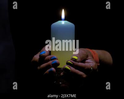Eine brennende Kerze in weiblichen Händen gehalten. Die Kerze hat einen bläulich-gelben Farbton. Die Nägel sind in den gleichen Farben lackiert. Stockfoto