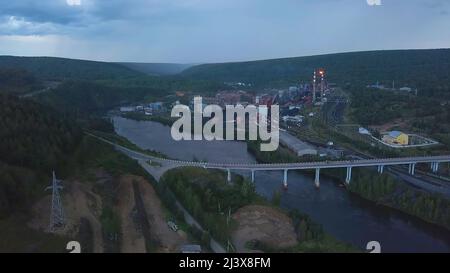 Eine Luftdrohnenlandschaft eines kleinen Dorfes mit Fluss und grünen bewaldeten Bergen. Clip. Eine Brücke über zwei Flussufer auf bewölktem Himmel Hintergrund Stockfoto