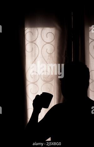 Ein erwachsener Mann schaut hinter den Vorhang, während er Kaffee trinkt. Konzept der Sperrung und Depression bei einer Pandemie. Stockfoto