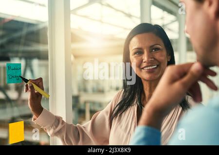 Ihre Vision zum Leben erwecken. Aufnahme einer selbstbewussten Geschäftsfrau, die ihrem Kollegen mit Haftnotizen an einer Glaswand im Büro eine Idee vorstellt. Stockfoto