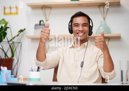 Ein junger Mann aus Asien hält die Daumen hoch, während er im Büro ein Callcenter-Headset trägt Stockfoto