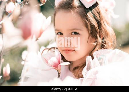 Schöne lächelnde Kind Mädchen 4-5 Jahre alt tragen stilvolle Prinzessin Kleid Geruch Magnolie Blume über blühenden Baum im Garten im Freien. Frühjahrssaison. Childho Stockfoto